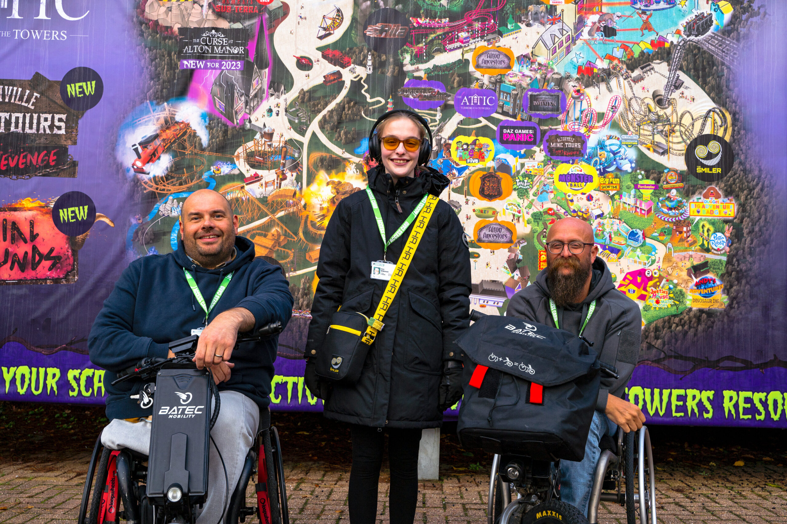 Three people, two men in wheelchairs, and a woman with tinted glasses and headphones, stood against a brightly colours theme park map, smiling